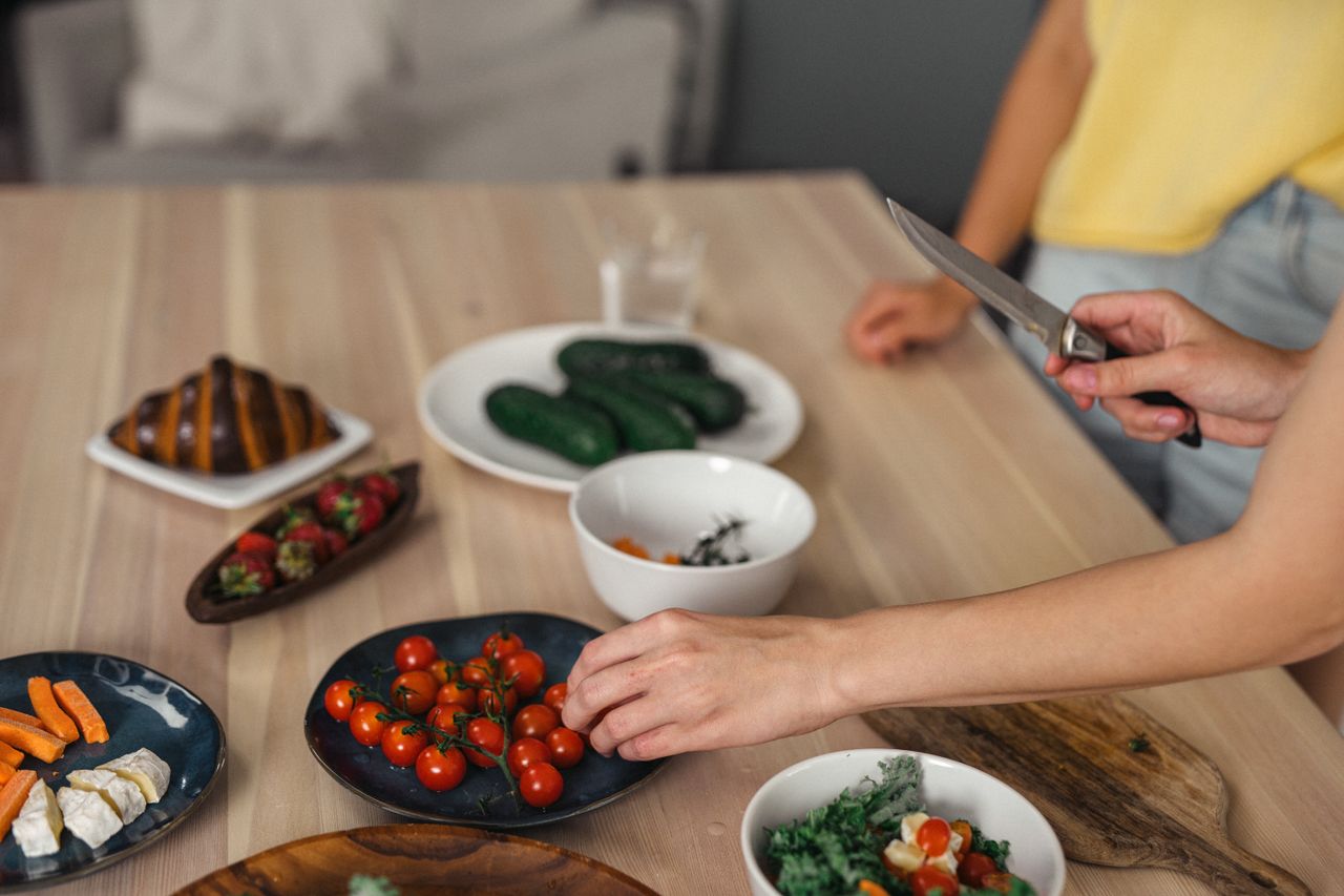Hands slicing and arranging fresh vegetables and fruits on wooden table indoors.